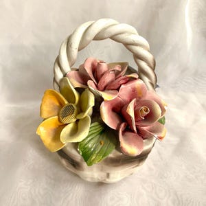 May include: A ceramic basket with a twisted handle, filled with colourful flowers. The flowers include yellow, pink, and red roses, with green leaves. The basket is off-white with brown accents.