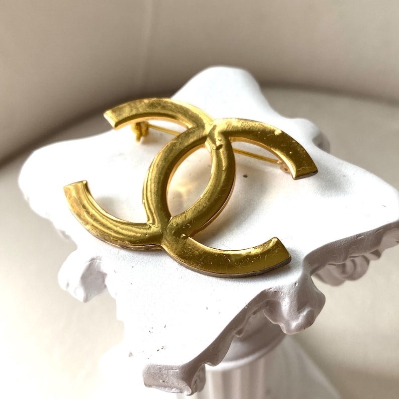 Pode incluir: Um broche de metal em tom dourado com o ic&oacute;nico log&oacute;tipo de dois C entrela&ccedil;ados. A superf&iacute;cie do broche apresenta um acabamento polido com sinais subtis de desgaste, incluindo pequenos riscos e ligeiras varia&ccedil;&otilde;es no brilho met&aacute;lico.