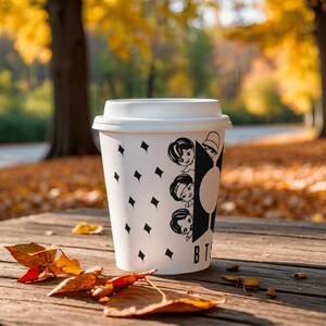 Pu&ograve; includere: Una tazza di caff&egrave; di carta bianca con coperchio, decorata con forme di diamanti neri e illustrazioni di personaggi dei cartoni animati. La tazza &egrave; su una superficie di legno con foglie cadute, con uno sfondo autunnale sfocato.