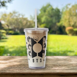 Pu&ograve; includere: Un bicchiere di plastica trasparente con una cannuccia bianca, riempito con una bevanda marrone chiaro. Il bicchiere presenta un design grafico nero con le lettere "BTS" e facce di cartoni animati. Lo sfondo &egrave; una scena sfocata all'aperto con erba verde e alberi.
