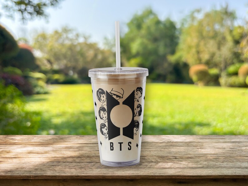 Pu&ograve; includere: Un bicchiere di plastica trasparente con una cannuccia bianca, riempito con una bevanda marrone chiaro. Il bicchiere presenta un design grafico nero con le lettere "BTS" e facce di cartoni animati. Lo sfondo &egrave; una scena sfocata all'aperto con erba verde e alberi.