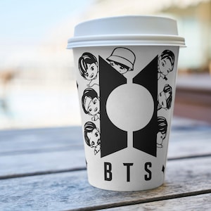Pu&ograve; includere: Una tazza di caff&egrave; di carta bianca con coperchio bianco, con un design grafico in bianco e nero. Il design include le lettere "BTS" e illustrazioni di cartoni animati di persone. La tazza &egrave; su un tavolo di legno.