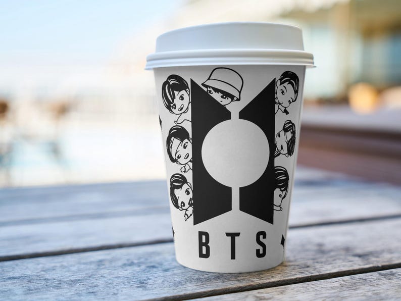 Pu&ograve; includere: Una tazza di caff&egrave; di carta bianca con coperchio bianco, con un design grafico in bianco e nero. Il design include le lettere "BTS" e illustrazioni di cartoni animati di persone. La tazza &egrave; su un tavolo di legno.
