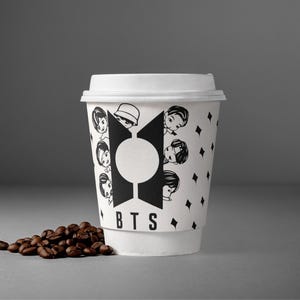 Pu&ograve; includere: Una tazza di caff&egrave; di carta bianca con un design in bianco e nero con facce di cartoni animati e le lettere "BTS". La tazza &egrave; piena di caff&egrave; e ha un coperchio bianco. Un piccolo mucchio di chicchi di caff&egrave; si trova accanto alla tazza.