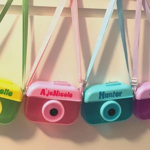 May include: Four colorful toy cameras with shoulder straps hang against a wall. Each camera is a different color: green and yellow, pink, teal, and purple. Each camera has a different name printed on it: Michelle, AJaNicole, Hunter, and Harley.