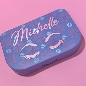 May include: A light blue contact lens case with a floral pattern and the name "Michelle" written in pink. The case has a design of two eyes with eyelashes.