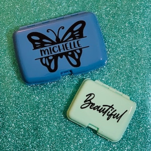 Puede incluir: Dos pequeñas cajas rectangulares con bisagras. La caja más grande es azul y presenta una mariposa negra con el nombre "Michelle" escrito en ella. La caja más pequeña es verde claro y tiene la palabra "Beautiful" escrita en cursiva negra.