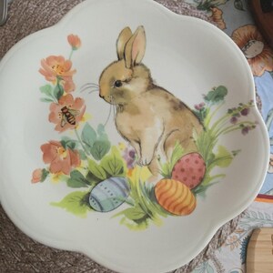 Może przedstawiać: Biały ceramiczny talerz z falistym brzegiem, ozdobiony centralną ilustracją brązowego królika w otoczeniu pomarańczowych kwiatów, małej pszczoły oraz trzech kolorowych, wzorzystych jaj wielkanocnych w odcieniach niebieskiego, pomarańczowego i różu w kropki.