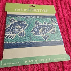 Peut inclure: Bordure décorative "playful spirit" avec deux poissons stylisés bleus et blancs sur un motif de vagues bleu et turquoise. L'emballage indique "restore & RESTYLE". La bordure mesure 13,3 cm de large.
