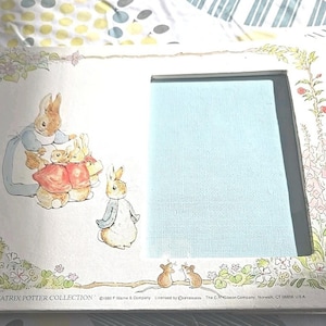 May include: A white picture frame featuring illustrations from The Beatrix Potter Collection. The frame has a rectangular opening with a light blue background. The frame is decorated with floral and bunny illustrations.