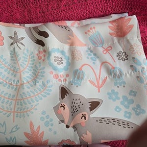 May include: A white fabric with a repeating pattern of a gray fox, flowers, and foliage in light blue and peach. The fox has a pink nose and ears. The design is likely for a curtain or other home decor item.