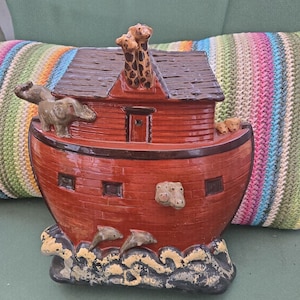 May include: A ceramic Noah's Ark figurine featuring a glossy reddish-brown boat with a dark brown roof. Two giraffes stand on the roof, while an elephant, two small animals, and two dolphins are sculpted onto the sides above dark waves.