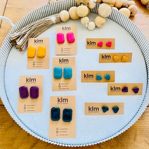 May include: Assortment of colorful geometric shaped earrings displayed on a tray. The earrings are made of polymer clay and come in a variety of colors, including teal, purple, pink, yellow, and green. The earrings are labeled with the brand name "klm designs".