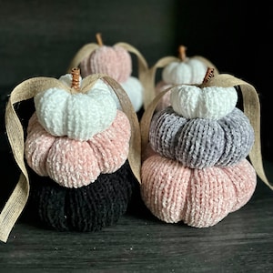 Op de afbeelding: Drie gehaakte pompoenen in tinten roze, wit en grijs. De pompoenen zijn op elkaar gestapeld en hebben een rustieke, handgemaakte uitstraling.