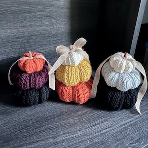 Op de afbeelding: Drie gebreide pompoenversieringen in verschillende kleuren. De pompoenen zijn op elkaar gestapeld en hebben een rustieke, herfstachtige uitstraling.