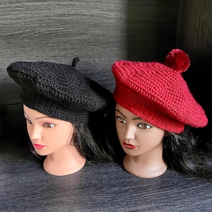 May include: Two crocheted berets, one black and one red, are worn by mannequins with long black hair. The berets are both styled with a slight tilt to the side.