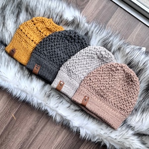 Puede incluir: Cuatro gorros de punto en diferentes colores, incluyendo amarillo mostaza, gris, beige y rosa. Cada gorro tiene una pequeña etiqueta de cuero con la letra "X" en ella.