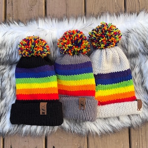 May include: Three rainbow striped knitted hats with pom poms. The hats are in different colours, with one being grey, one being white, and one being black.