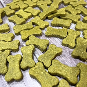 May include: A collection of bone-shaped dog treats in various sizes. The treats are a light green color and appear to be made of a textured, grainy material. They are scattered on a wooden surface.