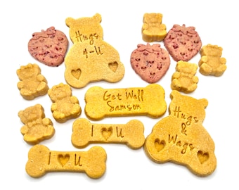 Op de afbeelding: Assortiment van hondensnacks in de vorm van botten en beren. De snacks zijn versierd met berichten zoals "Hugs 4-U", "Get Well Samson", "I Love You" en "Hugs & Wags".