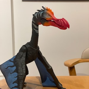 May include: A black and blue toy pterosaur with a red beak and orange accents. The toy has articulated wings and legs, and is posed on a wooden surface. The pterosaur's neck is striped with gray and white.