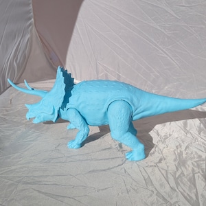 May include: A light blue plastic triceratops dinosaur toy. The toy has three horns, a frill, and a long tail. The dinosaur is standing on four legs and has a detailed texture. The toy is approximately 18 inches long.