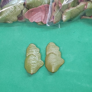 Puede incluir: Dos hojas de plástico verde con una superficie texturizada. Las hojas tienen forma de hoja tradicional con una punta puntiaguda y una base redondeada.