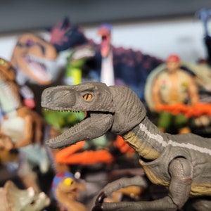 以下が含まれることがあります： 首に白いストライプのある灰色と茶色の恐竜のおもちゃのクローズアップ。恐竜は大きく開いた口と鋭い歯を持つ大きな頭を持っています。恐竜の目は明るいオレンジ色です。