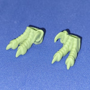 Puede incluir: Dos pies con garras de color verde claro, probablemente de una figura. Cada pie tiene tres dedos con garras afiladas y curvadas. Los pies están detallados con superficies texturizadas, lo que sugiere un origen reptiliano o aviar.