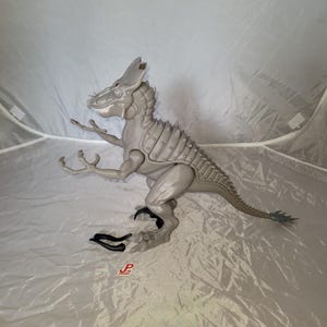 Könnte beinhalten: Ein graues Dinosaurier-Spielzeug mit beweglichen Gliedmaßen und einem detaillierten, strukturierten Design. Das Spielzeug hat einen stacheligen Rücken, einen langen Schwanz und einen Kopf mit Hörnern. Eine schwarze Klaue und ein Logo befinden sich in der Nähe des Dinosauriers.