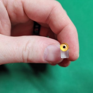 May include: A close-up of a yellow and black glass eye, mounted on a clear plastic base. The eye is used for taxidermy or craft projects.