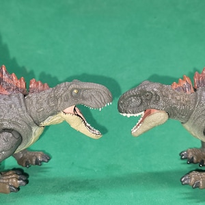 Puede incluir: Dos dinosaurios de juguete enfrentados. El dinosaurio de la izquierda es marrón y gris con una cresta roja y marrón. El dinosaurio de la derecha es gris con una cresta roja.