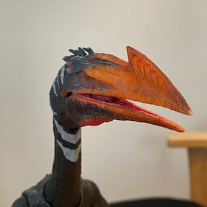 Puede incluir: Figura detallada de un Pteranodon, un reptil volador prehistórico. La figura presenta un pico anaranjado texturizado, una cabeza azul oscuro y negra con rayas blancas y un cuerpo oscuro. La boca está abierta, revelando un interior rojo.