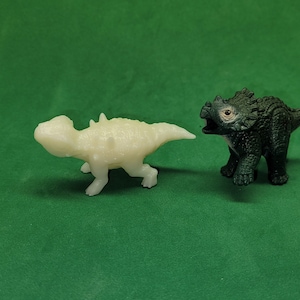 May include: Two small dinosaur figurines, one white and one black, on a green background. The white dinosaur is facing left and the black dinosaur is facing right.