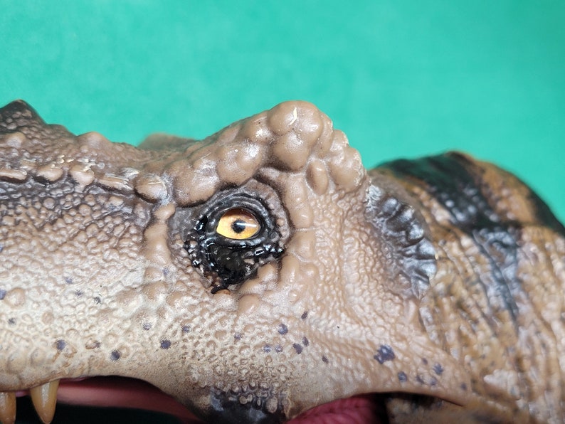 May include: Close-up of a dinosaur toy's eye. The eye is brown with a yellow iris and black pupil. The dinosaur's skin is textured and brown.