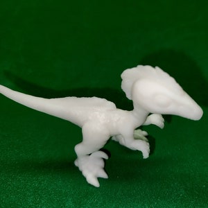 Może przedstawiać: Biała figurka dinozaura wydrukowana w 3D. Dinozaur jest mały i ma długi ogon oraz grzebieniową głowę.