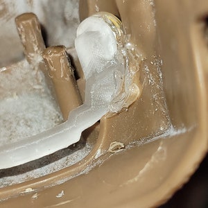 May include: Close-up of a clear plastic piece attached to a brown plastic surface with a white adhesive. The plastic piece is secured with a gold screw.