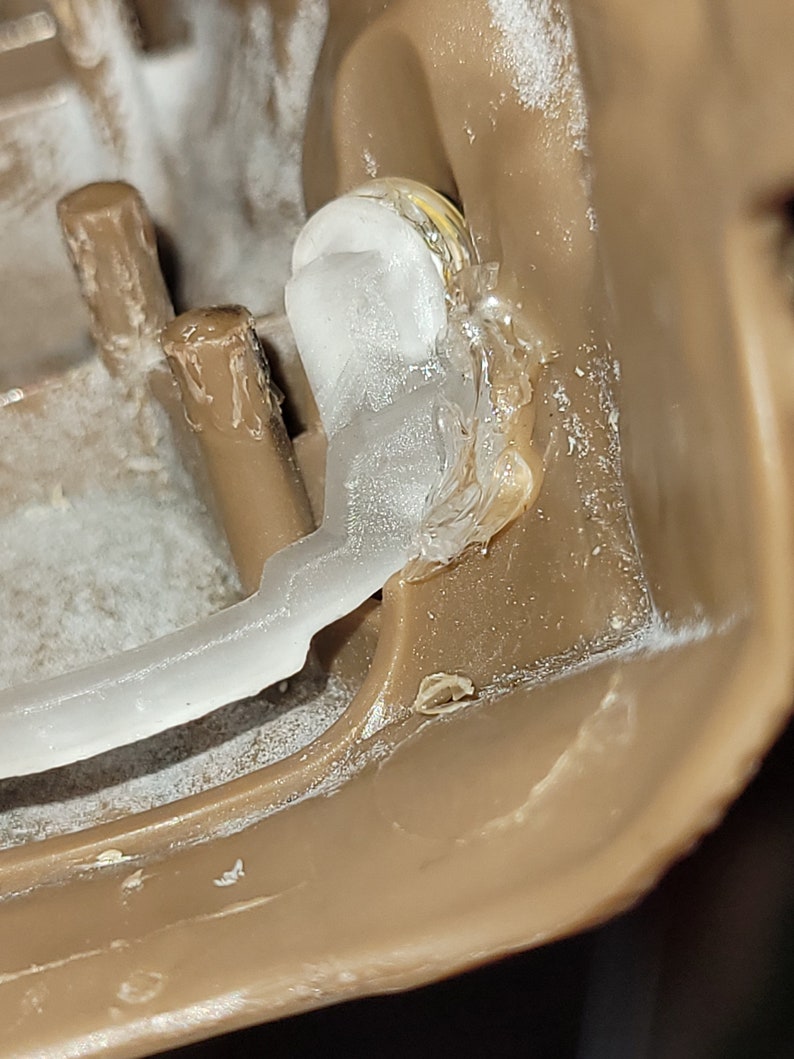 May include: Close-up of a clear plastic piece attached to a brown plastic surface with a white adhesive. The plastic piece is secured with a gold screw.