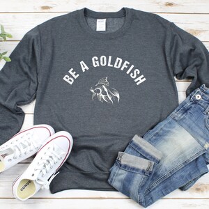 May include: A charcoal grey sweatshirt featuring the words "BE A GOLDFISH" in white, and a goldfish graphic. The image also shows blue jeans and white trainers with red detailing.