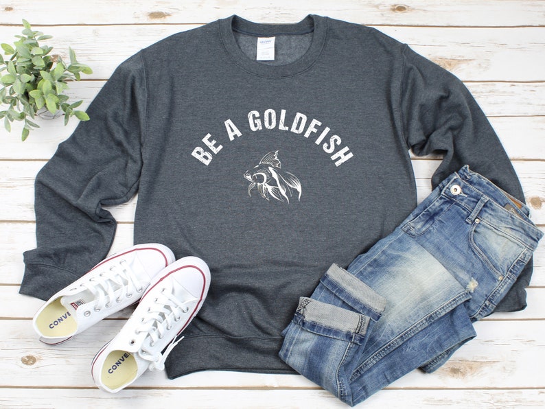 May include: A charcoal grey sweatshirt featuring the words "BE A GOLDFISH" in white, and a goldfish graphic. The image also shows blue jeans and white trainers with red detailing.