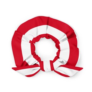 Puede incluir: Una scrunchie a rayas rojas y blancas con un lazo. La tela se recoge alrededor de una banda elástica central. La scrunchie está hecha de un material suave y es adecuada para varios peinados. El diseño es simple y llamativo.