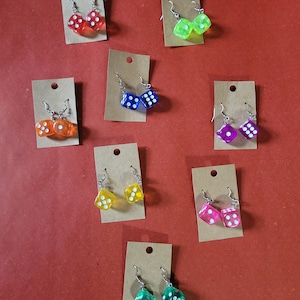May include: A collection of dice-shaped earrings in various colors, including red, green, yellow, pink, blue, and orange. Each pair of earrings is attached to a small, brown cardboard display card with a silver-colored hook.