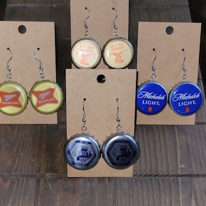 May include: A set of six pairs of earrings made from beer bottle caps. The earrings are silver and feature different beer brands, including Miller High Life, Michelob Light, and 21 to Enjoy.
