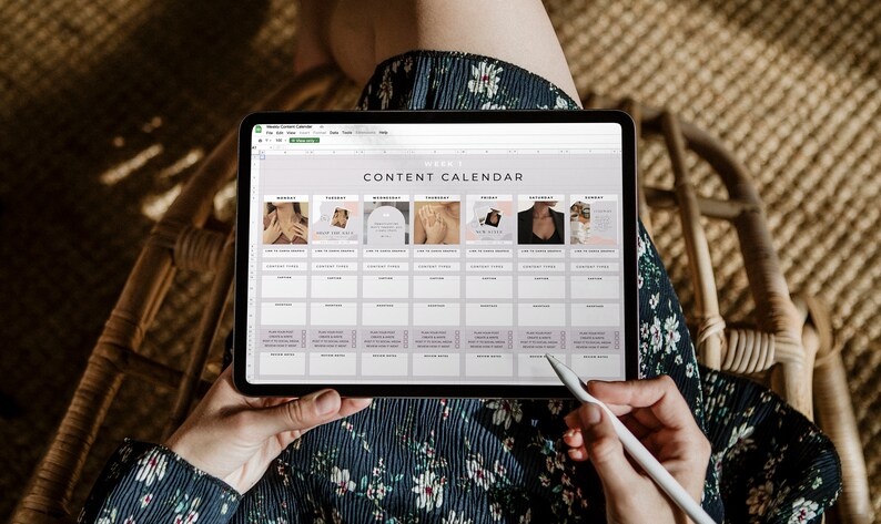 May include: A digital tablet displaying a "Content Calendar" spreadsheet. The calendar is organized in a grid format with sections for each day of the week, featuring images and text. A stylus is held near the screen, and the person is wearing a floral dress.