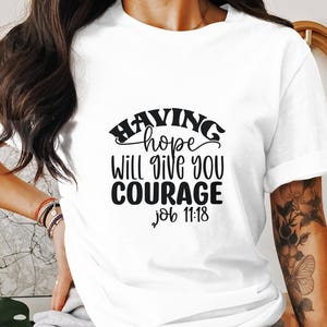 May include: White t-shirt with the text "HAVING hope WILL GIVE YOU COURAGE Job 11:18" in black. The shirt has short sleeves and is being worn by a person. The shirt is a casual wear item.