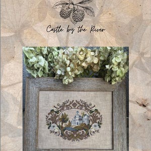 May include: Framed cross-stitch art titled "Castle by the River" by Blackberry Bay Designs. The artwork depicts a castle scene surrounded by floral and foliage embroidery. The frame is a light gray wood.
