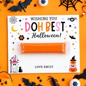 Puede incluir: Una tarjeta rectangular blanca con el texto WISHING YOU DOH BEST Halloween! y LOVE EMILY, decorada con iconos coloridos de Halloween que incluyen un fantasma, un gato y calabazas. Una barra de plastilina naranja está fijada en el centro.