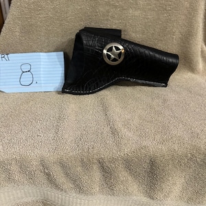 Op de afbeelding: Zwarte leren holster met een zilveren sterembleem. De holster heeft een textuurpatroon en een gebogen ontwerp. Het sterembleem is gecentreerd op de voorkant van de holster. De holster is ontworpen om een vuurwapen te bevatten.