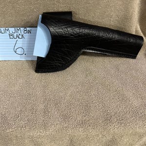 Op de afbeelding: Een zwarte leren schede, mogelijk voor een gereedschap of wapen, met een textuuroppervlak. De schede is ongeveer 20 cm lang. Een blauw en wit papier met de tekst "SLIM JIM 8 IN BLACK" is zichtbaar.