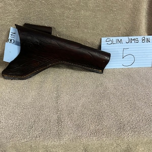 Op de afbeelding: Een donkerbruine leren holster met reliëfdetails. De holster wordt gepresenteerd op een neutraal gekleurd oppervlak. Een blauwe plakbriefje leest "SLIM JIMS 8IN" en het nummer "5".